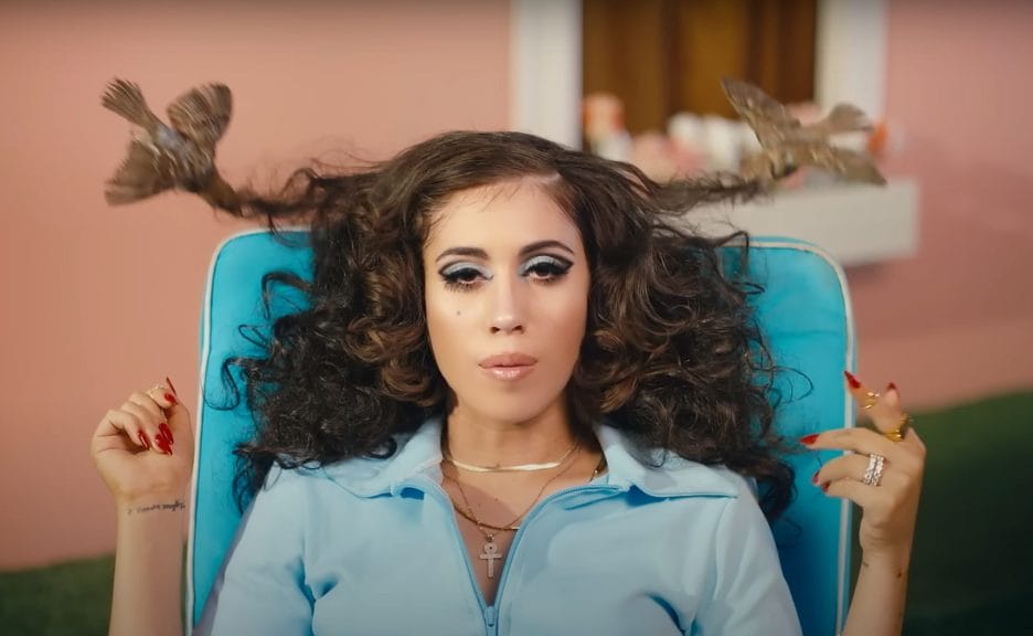 Kali Uchis is sitting in a lawn chair with curly hair, birds are pulling the singer's hair in different directions.